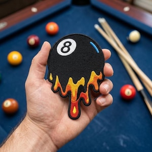 May include: An embroidered patch shaped like a black 8-ball with a white number eight, featuring a melting effect at the bottom in shades of yellow, orange, and red, with a small blue highlight on the upper right side of the circular ball.