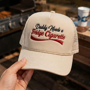 May include: A beige trucker hat featuring a mesh back and a structured front panel. The front displays black and red embroidered text that reads Daddy Needs a Fridge Cigarette, accented by a red swoosh design underneath the lettering.