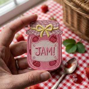 May include: An embroidered patch shaped like a glass jar filled with pink jam, featuring a yellow bow on the lid and a white label with the text JAM! in pink letters. The design includes small red strawberry illustrations on the jar body.