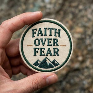 May include: A circular embroidered patch with a cream-colored background and a dark green border. The text FAITH OVER FEAR is stitched in dark green, centered above a dark green mountain range graphic. Two small horizontal gold lines flank the text.