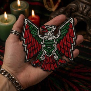 May include: An embroidered patch featuring a stylized eagle with wings spread wide. The design incorporates vibrant red, green, and white thread colors, with intricate geometric patterns outlining the bird's feathers, head, and talons in black.