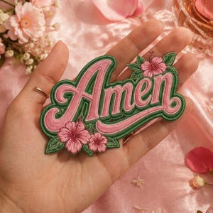 May include: An embroidered patch featuring the word Amen in pink script lettering with a dark green border. The design is decorated with pink hibiscus flowers and green leaves positioned at the top right and bottom left corners of the text.