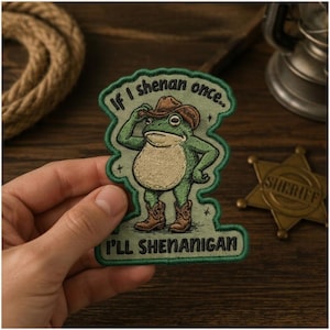 May include: An embroidered patch featuring a green frog wearing a brown cowboy hat and brown cowboy boots. The text reads, "If I shenan once.. I'LL SHENANIGAN" in black lettering. The patch has a green border and a light green background.
