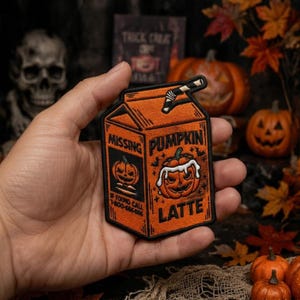 May include: An embroidered patch shaped like a milk carton in orange and black. The front features a jack-o'-lantern illustration with white icing, the text PUMPKIN LATTE, and a side panel with MISSING, a small pumpkin, and 1-800-666-666.