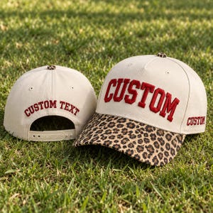 May include: Two off-white baseball caps with leopard print brims. The front cap features the word CUSTOM in red embroidery, while the back cap displays the text CUSTOM TEXT in red. Both caps have a small leopard print button on the crown.