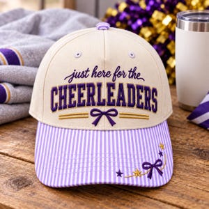 May include: A cream-colored baseball cap with a purple and gold embroidered text that reads "just here for the CHEERLEADERS". The cap features a purple and white pinstriped brim, a purple bow, and small embroidered stars on the right side.