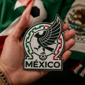 May include: A circular embroidered patch featuring a dark green eagle with spread wings perched on a soccer ball. The design includes a border with green and red accents and the word MÉXICO in dark green capital letters on a white background.