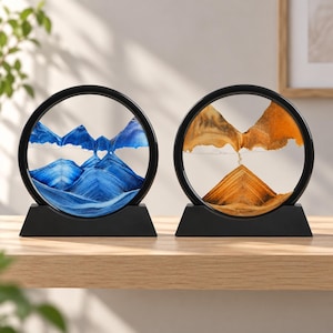 Könnte beinhalten: Zwei kreisförmige Sandkunst-Displays mit schwarzen Rahmen und Sockeln. Ein Display zeigt geschichteten blauen und weißen Sand in Bergform, während das andere orangefarbenen, gelben und schwarzen Sand enthält, der eine ähnliche Landschaft bildet.