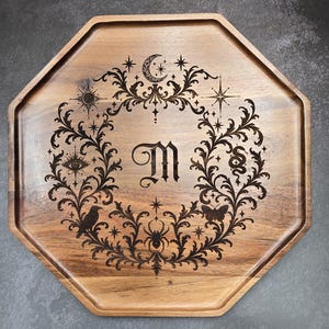 May include: An octagonal wooden tray featuring a laser-engraved design of a central letter M surrounded by a circular wreath of foliage, stars, a crescent moon, a sun, an eye, a snake, a spider, a butterfly, and a bird silhouette.