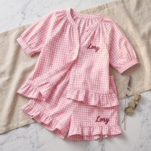 May include: A pink and white gingham pyjama set featuring a button-down top with three-quarter sleeves and a ruffled hem, paired with matching ruffled shorts. The name Lory is embroidered in dark red script on both the top and the shorts.