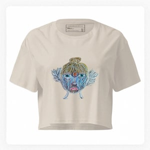 Puede incluir: Top corto color crema con una colorida ilustración de un rostro en tonos azules, rojos y marrones. La obra de arte incluye un diseño central en forma de ojo y elementos en forma de hojas. La camiseta tiene cuello redondo y mangas cortas.