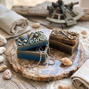 Könnte beinhalten: Zwei rechteckige Dekoboxen mit Deckel, eine blau und eine bronzefarben, mit Reliefverzierungen aus Muscheln, Seesternen und einem Steuerrad. Eine silberfarbene Perlenkette hängt aus der offenen blauen Box, die auf einer polierten Holzscheibe steht.