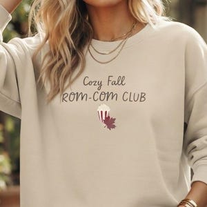 May include: A beige crewneck sweatshirt featuring the dark brown text Cozy Fall ROM-COM CLUB printed on the chest. Below the text is a small graphic illustration of a red and white striped popcorn bucket next to a single dark red maple leaf.