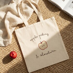 May include: A natural-colored canvas tote bag featuring two fabric handles and a graphic print. The design includes the text "apple picking & slowburn" in a dark brown script font, accompanied by a simple illustration of a sliced red apple.