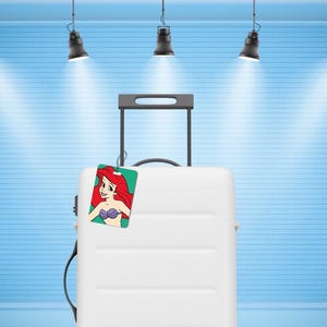 May include: A white hard-shell rolling suitcase with a retractable black handle and a rectangular luggage tag attached. The tag features a colourful illustration of a character with long red hair and a purple seashell top against a green background.