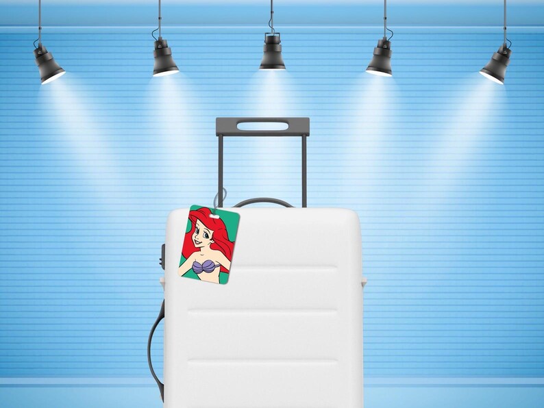 May include: A white hard-shell rolling suitcase with a retractable black handle and a rectangular luggage tag attached. The tag features a colourful illustration of a character with long red hair and a purple seashell top against a green background.