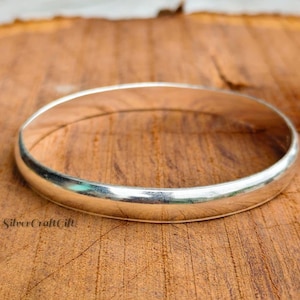May include: A smooth, polished silver-toned bangle bracelet with a rounded profile and a continuous circular band. The metal surface reflects light, creating bright highlights along the curve of the jewellery piece. The text SilverCraftGift is visible.