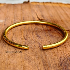 May include: A polished gold-tone metal cuff bracelet with a smooth, rounded surface and an open gap design. The bracelet features a simple, continuous circular shape with blunt, finished ends, showcasing a reflective, metallic yellow finish.