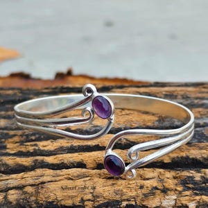 Può includere: Un bracciale rigido in argento dal design aperto con due estremità a spirale, ciascuna incastonata con una pietra preziosa di ametista viola, liscia e ovale. Il metallo ha una finitura lucida e il testo SilverCraftCilt è visibile in basso.