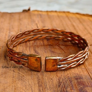 Op de afbeelding: Een koperen manchetarmband met een gevlochten ontwerp van meerdere gedraaide metalen strengen. De armband heeft aan beide uiteinden een brede, platte, rechthoekige sluiting, wat zorgt voor een open stijl met een metallic roodbruine afwerking.