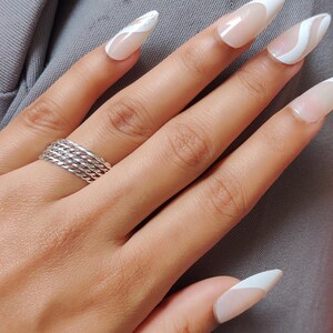 Könnte beinhalten: Eine Hand mit langen, mandelförmigen Nägeln. Die Nägel sind in einem weißen und nudefarbenen Farbschema lackiert, mit French-Tip-Design und weißen Wellenakzenten. Ein silberner Ring mit mehreren Bändern ziert den Ringfinger.