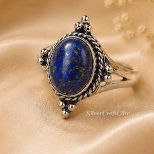 Op de afbeelding: Een zilveren ring met een ovale blauwe lapis lazuli edelsteen met gouden spikkels. De zetting heeft een gedraaide touwrand en kleine zilveren kraalaccenten aan de boven- en onderkant. De ringband is gespleten in twee delen bij de steen.