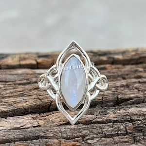 Puede incluir: Un anillo de plata con una piedra lunar central de corte marquesa con un aspecto translúcido e iridiscente. La banda de metal presenta un intrincado diseño de filigrana calada que enmarca la piedra en un patrón geométrico y simétrico.