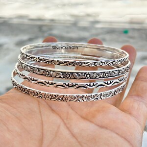 Puede incluir: Un conjunto de cuatro brazaletes de metal plateado con intrincados patrones en relieve. Los diseños incluyen motivos florales repetitivos, líneas onduladas y pequeñas formas de conchas. El texto SilverCraftGift es visible en la superficie.