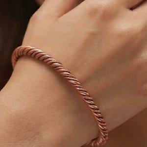 May include: A copper-colored bracelet featuring a twisted rope design. The metal band has a continuous spiral texture that wraps around the entire circumference, creating a uniform and detailed appearance throughout the piece of jewelry.