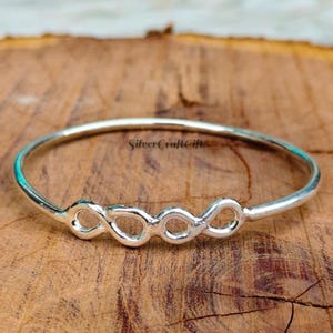 Może przedstawiać: Polerowana srebrna bransoletka typu bangle z centralnym motywem dwóch połączonych symboli nieskończoności. Metal ma gładkie, odblaskowe wykończenie, a napis SilverCraftGift jest subtelnie wygrawerowany na powierzchni metalowej obręczy.
