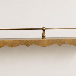 Op de afbeelding: Een wandplank met een metalen antieke messing afwerking. De plank heeft een geschulpte voorrand en een horizontale reling die wordt ondersteund door drie verticale stijlen. Het bevat twee zijdelingse montagebeugels met ronde gaten.