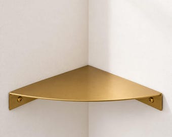 May include: A gold-colored metal corner shelf with a smooth, matte finish. The shelf features a triangular shape designed to fit into a corner, with two flat mounting brackets on each side, each secured by a single visible screw head.