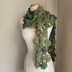 Könnte beinhalten: Ein handgefertigter, grüner Häkelschal, der auf einer Schaufensterpuppe präsentiert wird. Der Schal hat ein florales Design mit verschiedenen Grüntönen, wodurch ein strukturiertes und detailliertes Accessoire entsteht. Der Schal ist drapiert, um seine Länge und sein kompliziertes Muster zu zeigen.