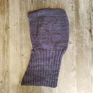 May include: A knitted balaclava in shades of blue and purple. The head covering features a ribbed neck and a hood that covers the head and neck. The balaclava is made of a soft, textured yarn.