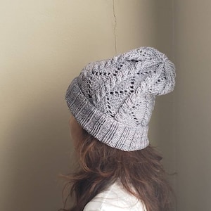 以下が含まれることがあります： ケーブル編みのパターンとレースのデザインが施されたグレーのニット帽。帽子は人の頭にかけられています。