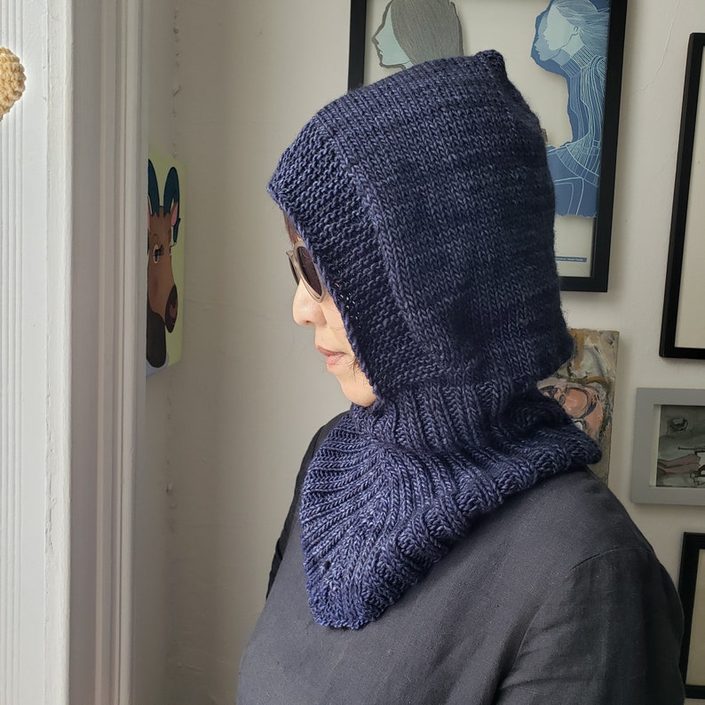 May include: A navy blue knitted hood with a ribbed neck and a pointed top. The hood is made of a thick, textured yarn and is worn by a person wearing sunglasses.