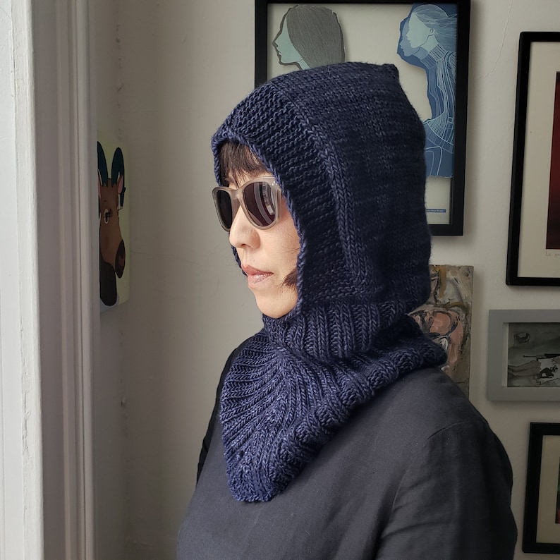 May include: A navy blue knitted hood with a cowl neck. The hood is made of a thick, textured yarn and features a pointed top. The cowl neck has a ribbed design. The person is wearing sunglasses and a black shirt.