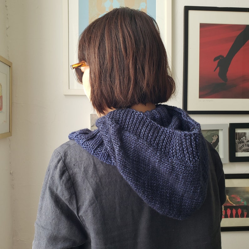May include: A navy blue knitted hood is worn over a gray shirt. The hood is made of a textured yarn and has a ribbed design. The person is wearing sunglasses and is facing away from the camera.