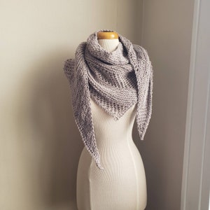May include: A light brown knitted triangular shawl draped over a dress form. The shawl is made with a simple stitch pattern and has a delicate, flowing drape.