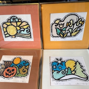 May include: A set of four framed square prints representing the seasons. Each print features bold black outlines and colored shapes. The top left shows a sun and star, top right shows flowers, bottom left shows a pumpkin, and bottom right shows snowflakes and trees.