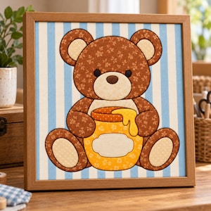 Op de afbeelding: Een ingelijst stoffen wandkunstwerk met een bruine teddybeer met bloemenpatroon die een gele honingpot met druipende honing vasthoudt. De beer staat tegen een achtergrond van verticale lichtblauwe en witte strepen in een houten lijst.