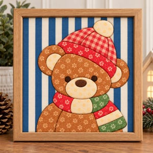Pode incluir: Uma peça de arte em tecido emoldurada apresentando um urso de peluche castanho com padrão floral, usando um gorro de inverno xadrez vermelho com pompom e um cachecol às riscas vermelhas e verdes. O urso está sobre um fundo de riscas verticais azuis e brancas.