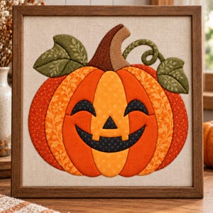 Può includere: Un'opera d'arte da parete quadrata incorniciata che presenta un'applicazione in tessuto a forma di zucca di Halloween. La zucca è composta da segmenti di tessuto arancione e giallo a motivi, con un gambo marrone scuro, foglie verdi e un volto nero sorridente.