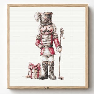 May include: A cross-stitch pattern featuring a nutcracker soldier in a red and white uniform with a tall hat, holding a staff. Beside the soldier is a gift box wrapped with a red bow. The design is framed in a light wood square frame.