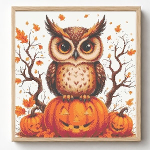 May include: A framed cross-stitch embroidery featuring a brown owl with large eyes perched on a carved orange jack-o'-lantern. The scene includes two smaller pumpkins and bare tree branches with scattered orange autumn leaves on a white background.
