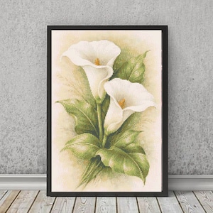 May include: A framed cross-stitch embroidery featuring two white calla lilies with yellow centers and green leaves. The artwork is set against a soft, cream-colored background with a subtle grid pattern, enclosed in a simple black rectangular frame.