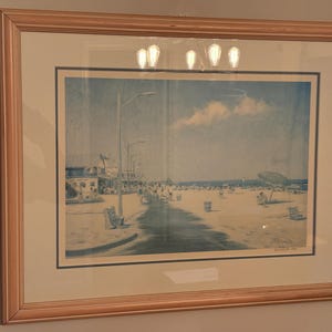 May include: A framed print featuring a blue-toned beach scene with a boardwalk, street lamps, and beachgoers under a cloudy sky. The artwork is enclosed in a light-colored wooden frame with a white mat and a thin dark blue inner border.