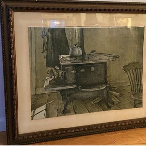 Puede incluir: Una impresión enmarcada que muestra una ilustración en blanco y negro de una estufa de leña de hierro fundido antigua con una tetera encima. La estufa se asienta sobre un suelo de madera junto a una silla, con un abrigo colgado en la pared.