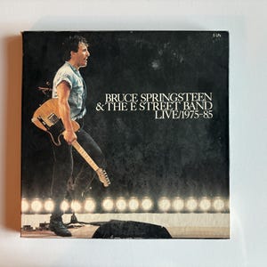 Puede incluir: Una caja cuadrada de discos de vinilo titulada Bruce Springsteen & The E Street Band Live/1975-85. La portada presenta un fondo negro con una fotografía de un músico tocando una guitarra eléctrica de color claro en un escenario con luces.