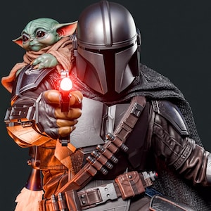 A detailed statue of a character from the Star Wars franchise, holding a gun and pointing it at a smaller figure, likely Baby Yoda, who is wearing a helmet.